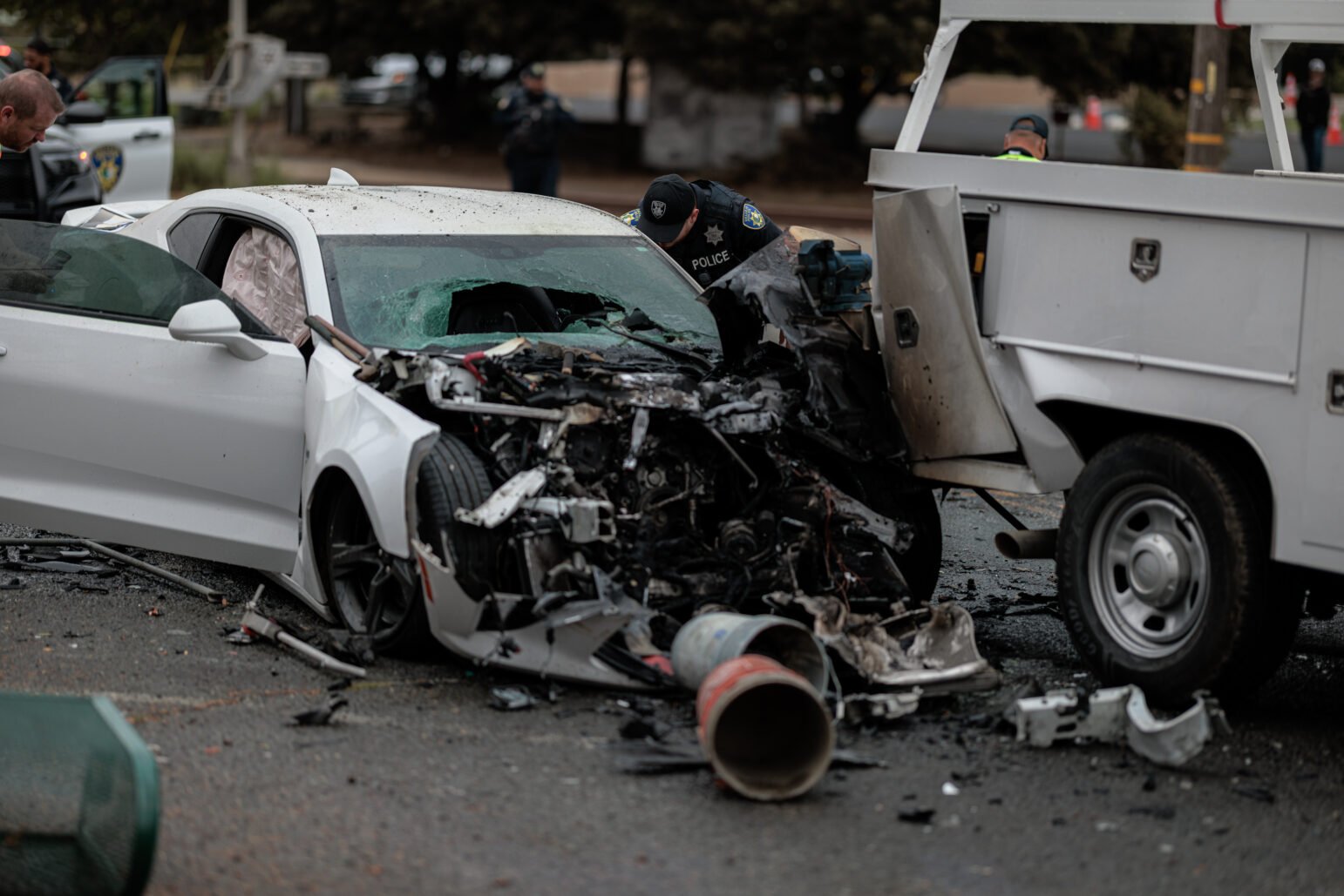 A white Chevrolet Camaro with severe front-end damage is seen after a collision with a utility truck. The car’s windshield is shattered, and airbags have deployed. A police officer inspects the wreckage, with debris scattered around the scene.