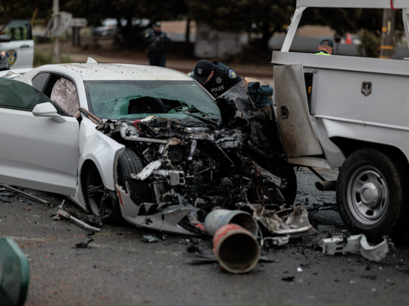 A white Chevrolet Camaro with severe front-end damage is seen after a collision with a utility truck. The car’s windshield is shattered, and airbags have deployed. A police officer inspects the wreckage, with debris scattered around the scene.