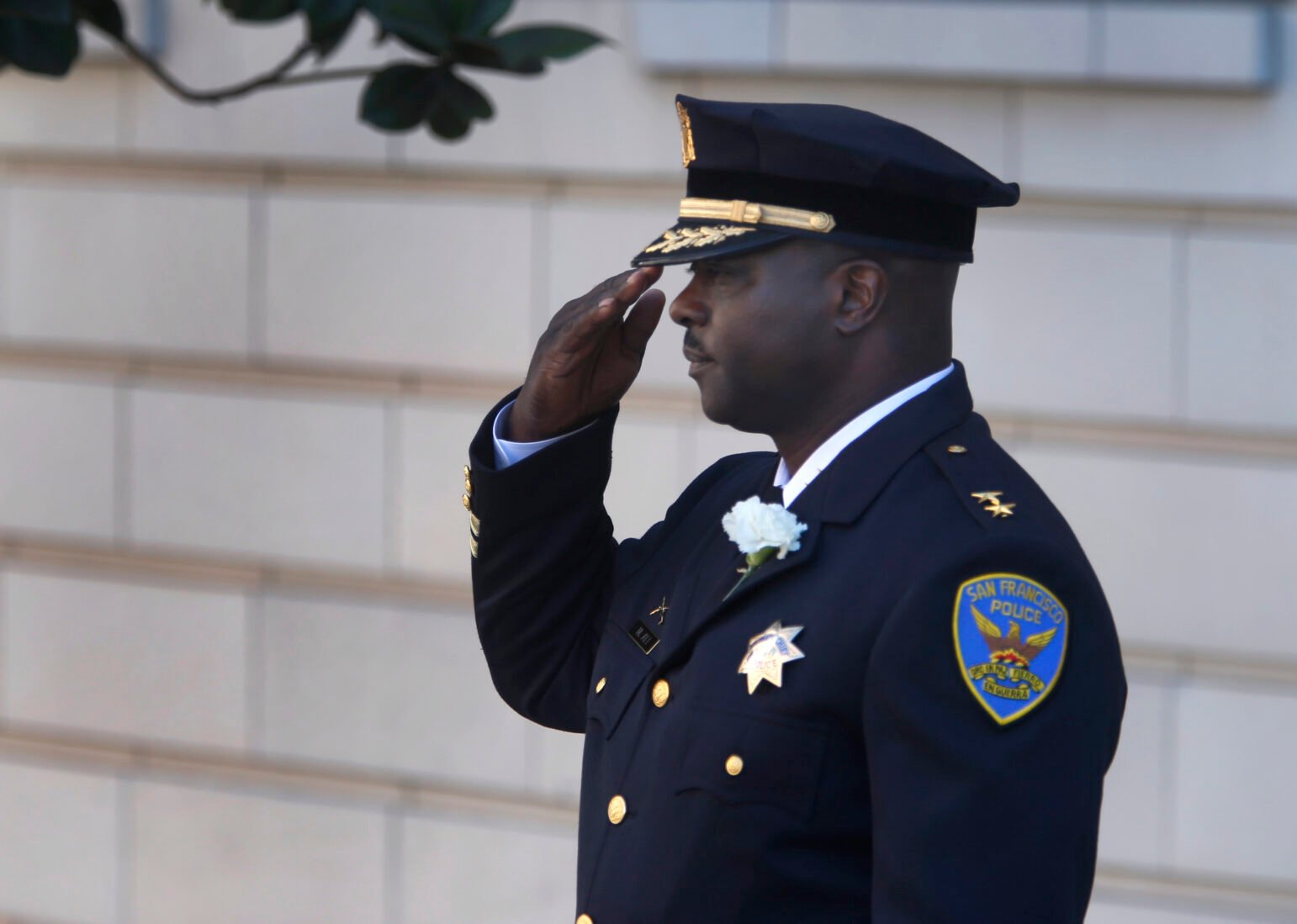 A police officer, dressed in formal uniform with gold accents and a badge, salutes while standing outdoors. The officer wears a San Francisco Police Department patch on his shoulder and a white flower pinned to his chest. His face is solemn, and he stands in profile against a light-colored building in the background.