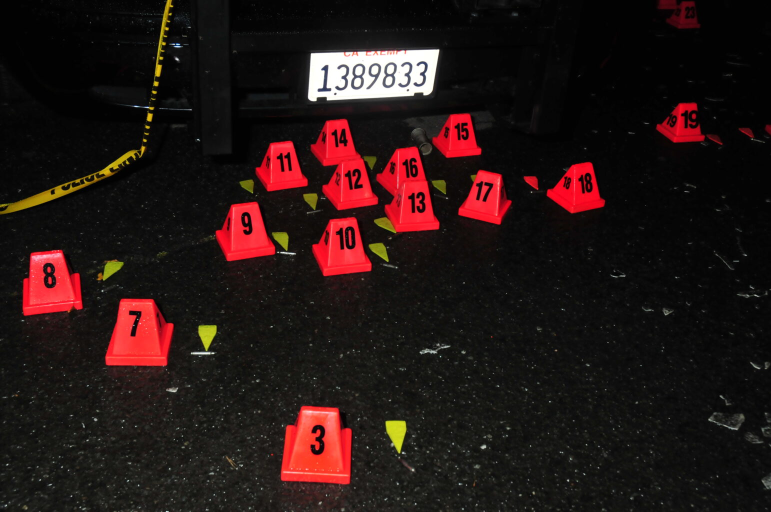 A series of bright orange evidence markers with black numbers are placed on a dark, wet road, indicating evidence at a crime scene. The markers are arranged near a vehicle with a visible license plate, and pieces of debris or evidence are scattered around the markers. Yellow police tape is partially visible, draped near the markers.