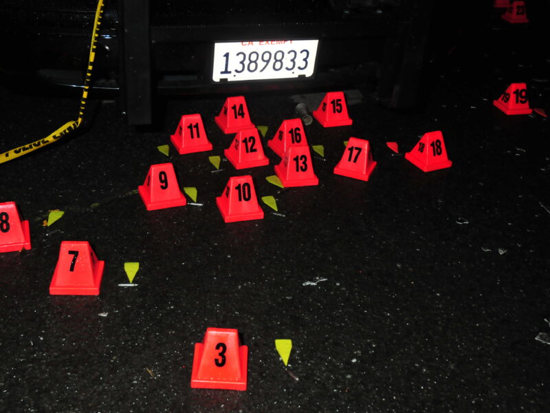 A series of bright orange evidence markers with black numbers are placed on a dark, wet road, indicating evidence at a crime scene. The markers are arranged near a vehicle with a visible license plate, and pieces of debris or evidence are scattered around the markers. Yellow police tape is partially visible, draped near the markers.