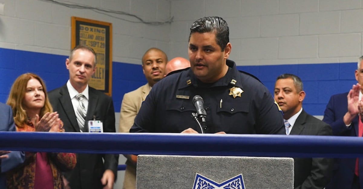 A police officer, wearing a uniform with a badge, speaks at a podium in front of a small crowd. Several individuals, including city officials and colleagues, stand behind him. The event appears to be held in a community or training center, with a boxing poster in the background.