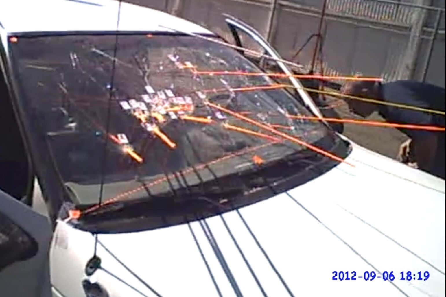 Photograph of the windshield of a white car showing several trajectory bars and signs indicating the direction of shots fired into the vehicle. The photo from September 6, 2012 shows an investigator leaning over to the passenger side and examining the crime scene.