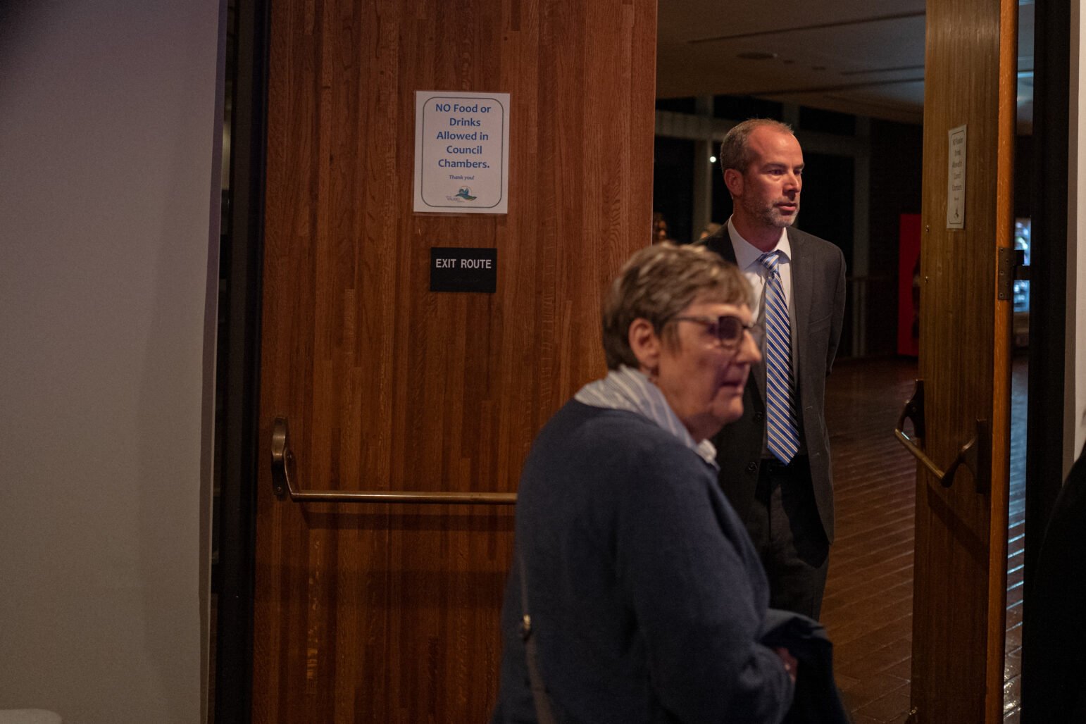 A man in a suit stands by a doorway marked "Exit Route" in the Vallejo City Council Chambers, while a woman walks past in the foreground.