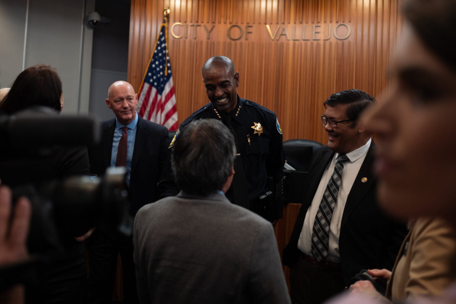 A Chief of Police and city officials gather, smiling and conversing under the "City of Vallejo" sign, with the American flag in the background.