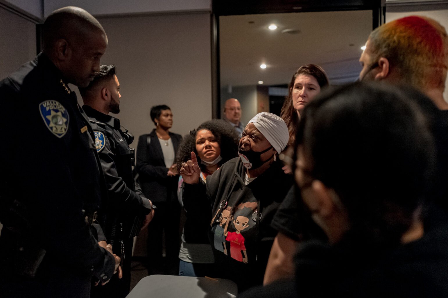 A woman wearing a headscarf and a graphic t-shirt stands in a heated exchange with two uniformed Vallejo police officers. Her finger is raised as she speaks, with other individuals, including another woman and a man, visible in the background observing the interaction. The scene is tense, with the woman’s facial expression showing frustration.