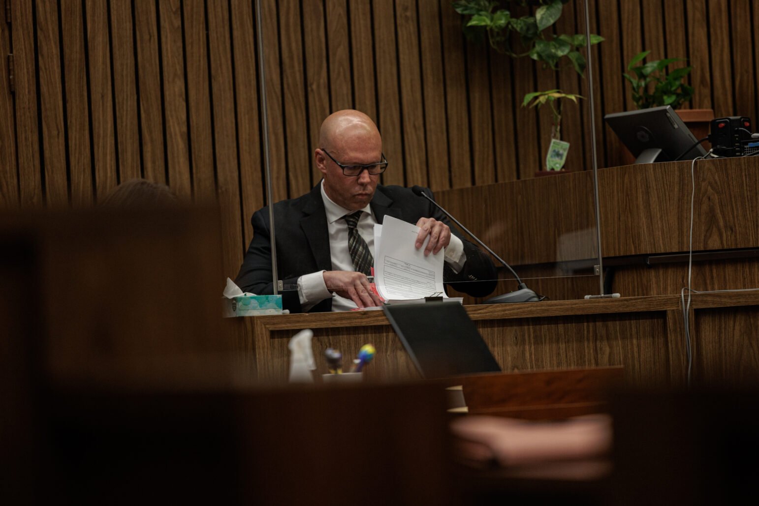 A bald man in a dark suit and tie sits at a wooden courtroom witness stand, carefully flipping through a stack of papers. He wears glasses and appears focused as he prepares documents, with courtroom elements such as a microphone, wood paneling, and small plants visible around him.