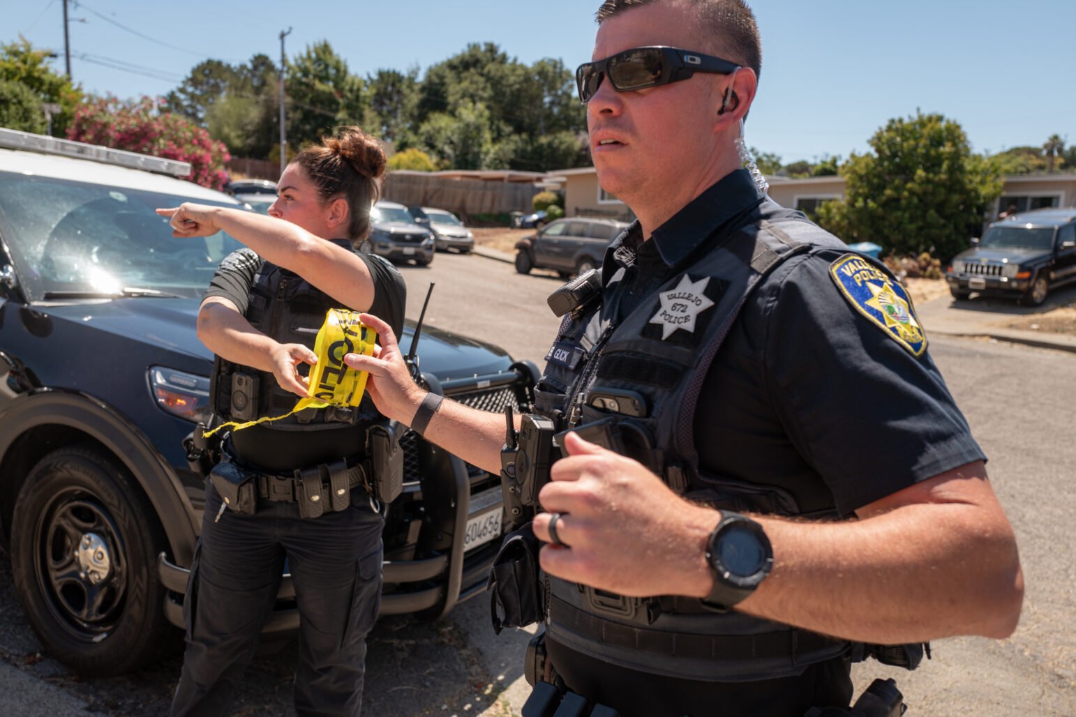 Two Vallejo police officers are seen at a crime scene. One officer holds yellow police tape and gestures, while the other officer, wearing sunglasses, looks on with a serious expression.