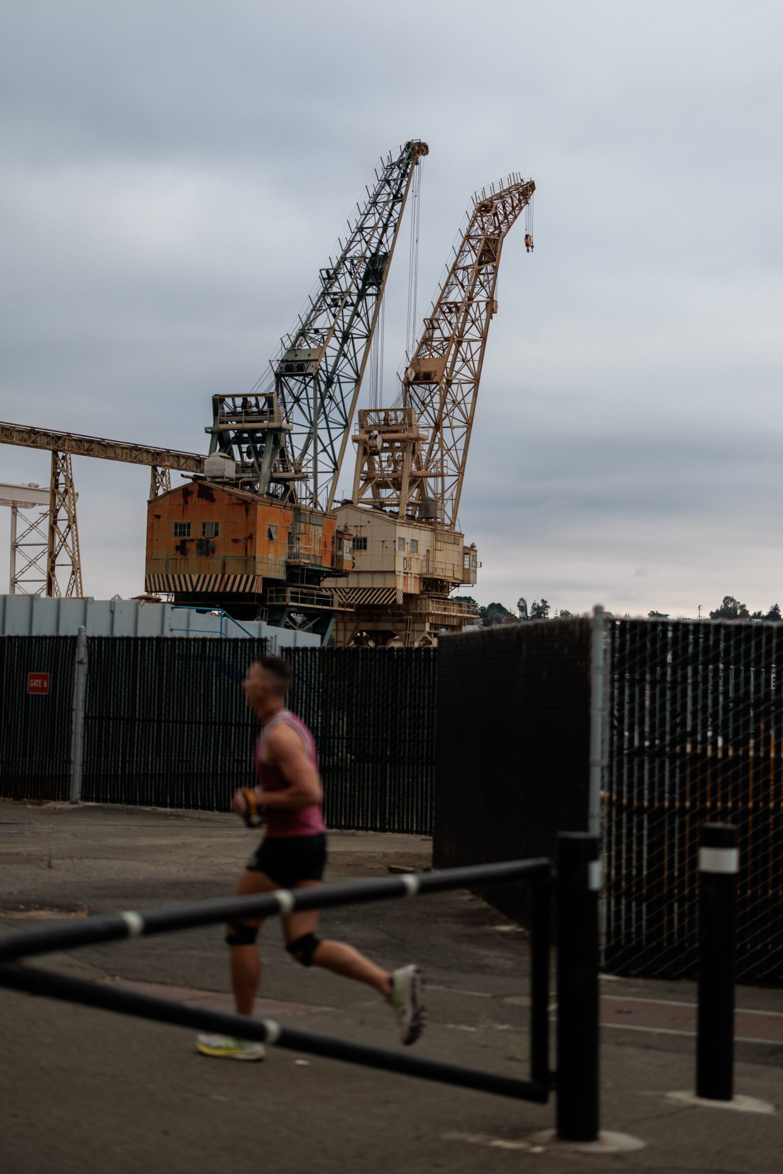 A jogger passes by tall, rusted cranes on Mare Island, with a cloudy sky above and a chain-link fence in the foreground.