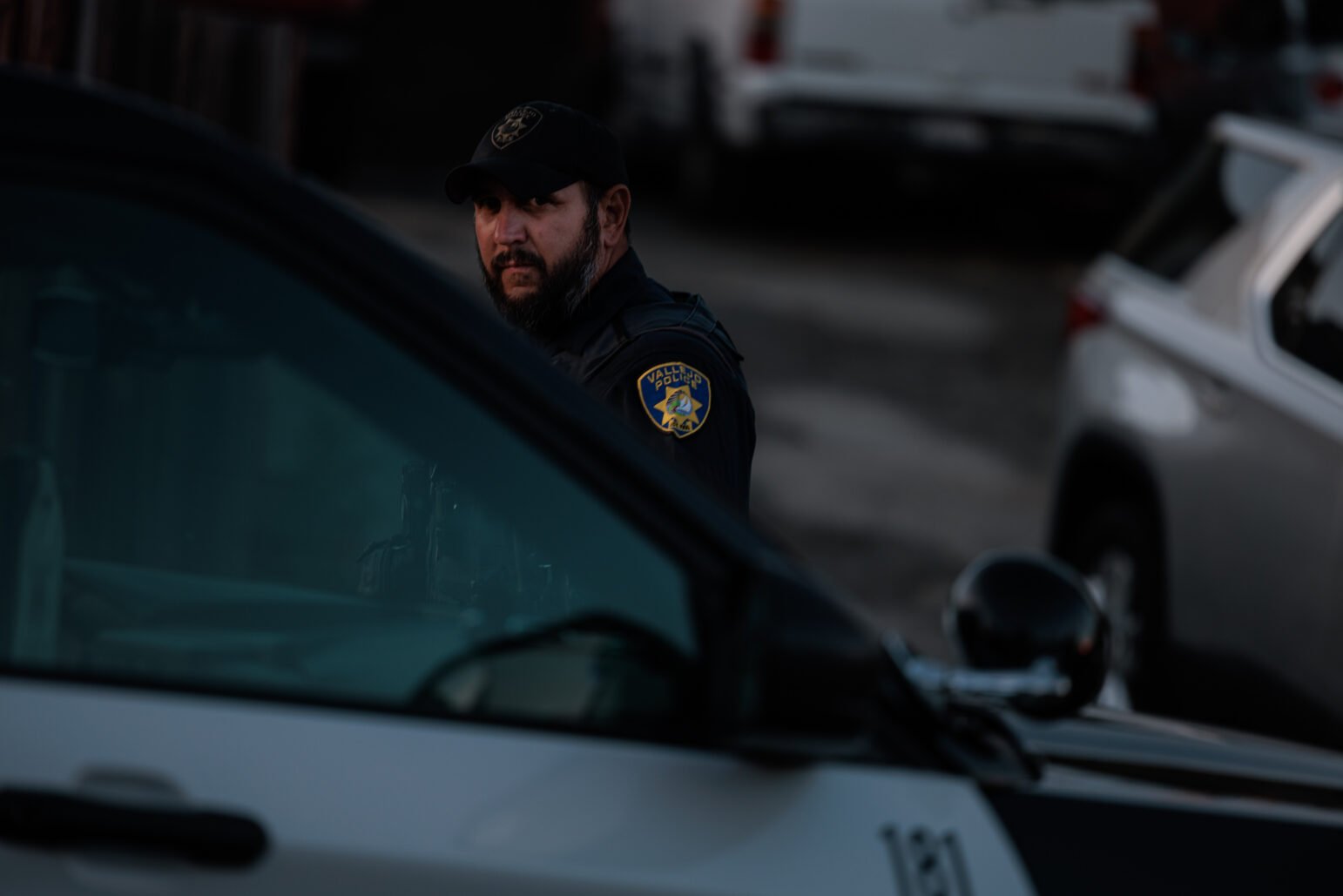 A Vallejo police officer, wearing a black cap, peers from behind a police vehicle, focused and alert.