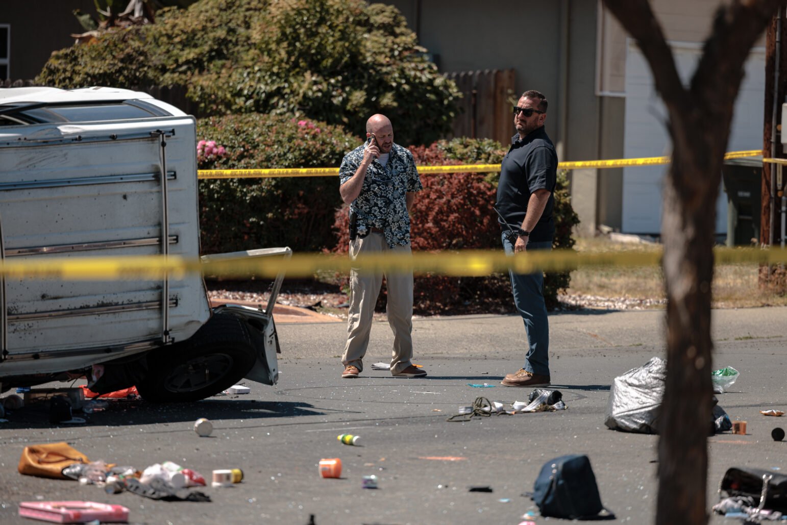 Two men stand behind yellow police tape in a residential area. They survey the scene where a small white vehicle is overturned, with debris scattered on the road.