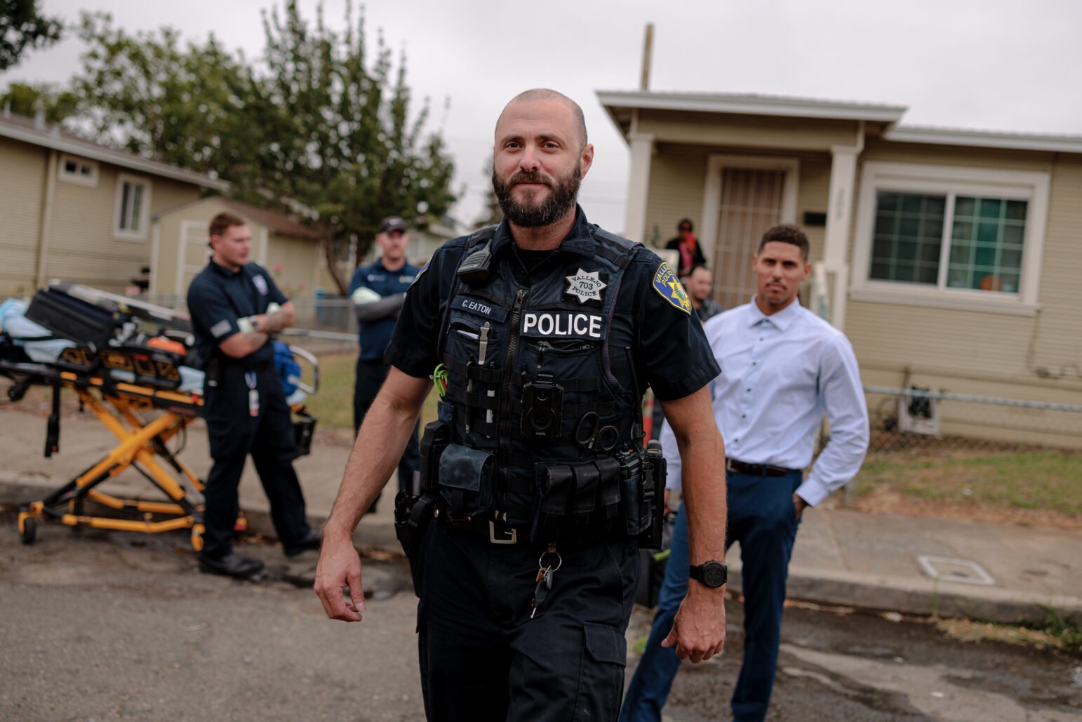 A Vallejo police officer walks confidently toward the camera. In the background, emergency personnel and a stretcher are visible.