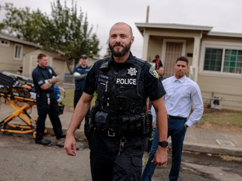 A Vallejo police officer walks confidently toward the camera. In the background, emergency personnel and a stretcher are visible.