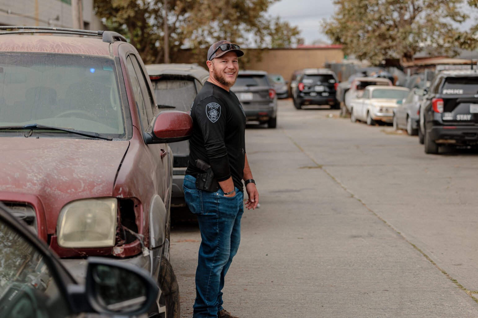 A Vallejo police officer, dressed in casual attire, stands near a red SUV in a parking area, smiling and relaxed. Several police vehicles are parked in the background.