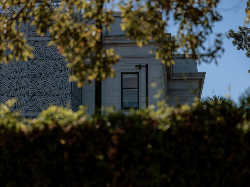 A partial view of the Old Solano Courthouse, framed by leafy trees and a hedge in the foreground. The building's light gray stone exterior is partially obscured, with modern patterned metalwork visible on one side. A single window is seen through the foliage, hinting at the structure’s historical architecture.