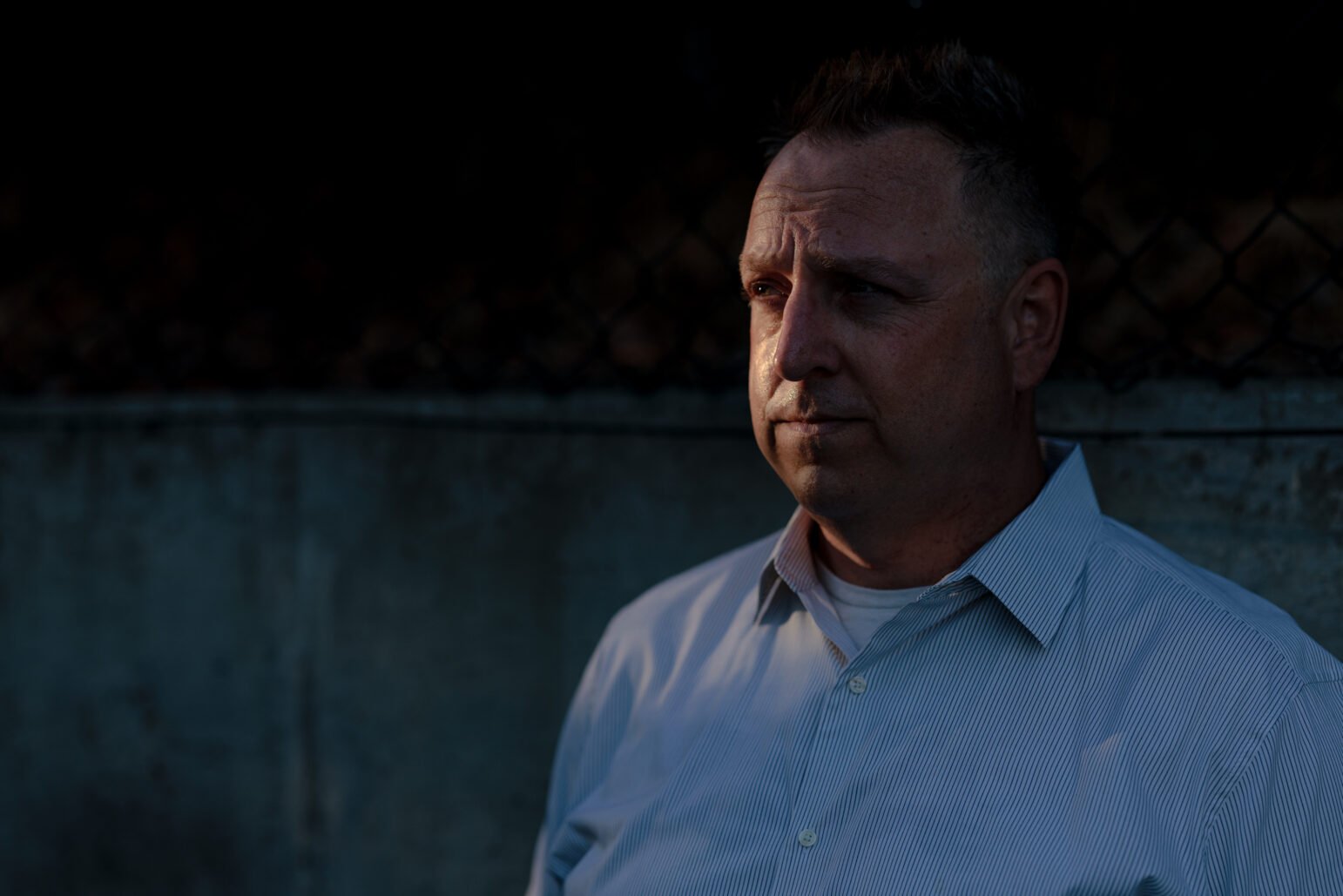 A man stands in front of a dimly lit concrete wall, gazing into the distance with a contemplative expression.