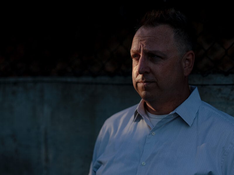 A man stands in front of a dimly lit concrete wall, gazing into the distance with a contemplative expression.