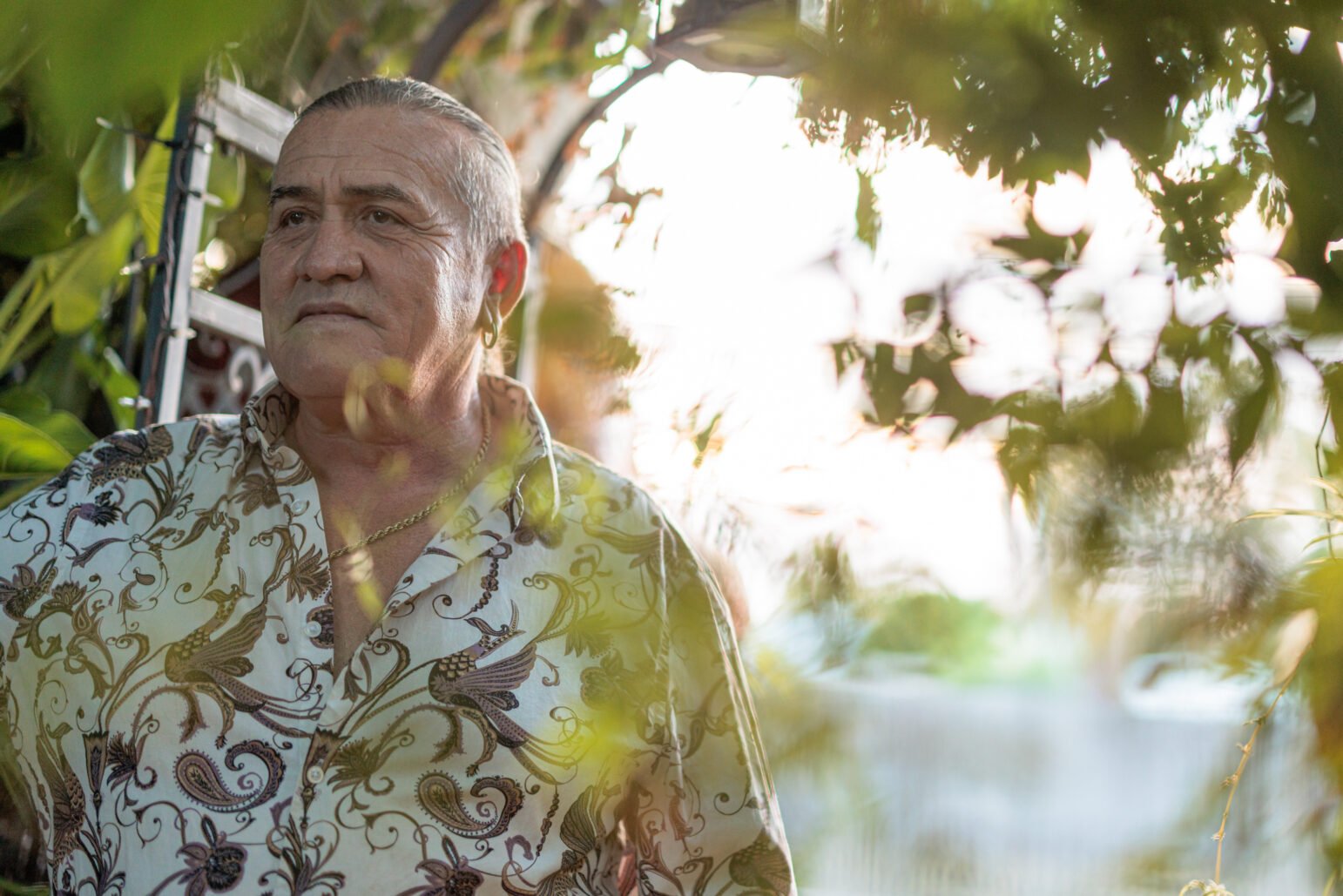 Mario Saucedo gazes to the side through a frame of green foliage. He wears a patterned shirt with gold jewelry and is illuminated by soft, natural light in an outdoor garden setting.