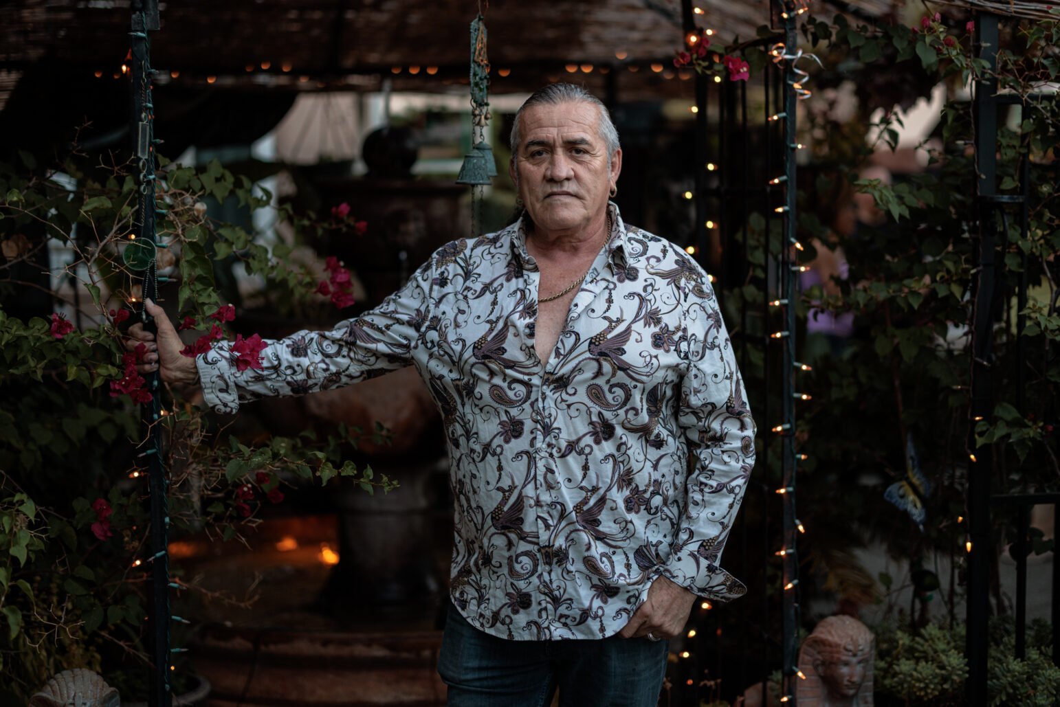 Mario Saucedo, dressed in a patterned shirt, stands with one hand holding a trellis adorned with string lights and pink flowers, looking directly at the camera. The setting is in a garden during dusk.