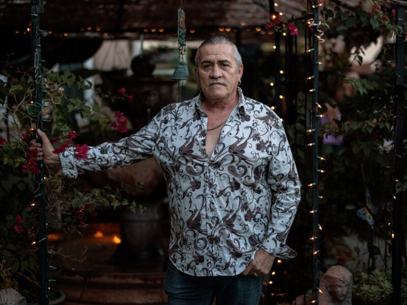 Mario Saucedo, dressed in a patterned shirt, stands with one hand holding a trellis adorned with string lights and pink flowers, looking directly at the camera. The setting is in a garden during dusk.