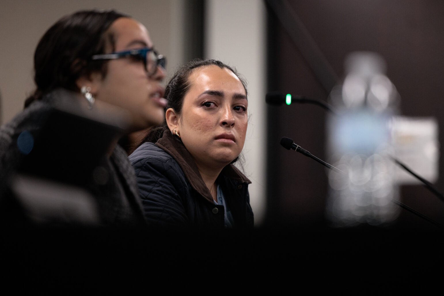 A woman sitting at a microphone during a public hearing, looking emotional, with another person speaking in the foreground.