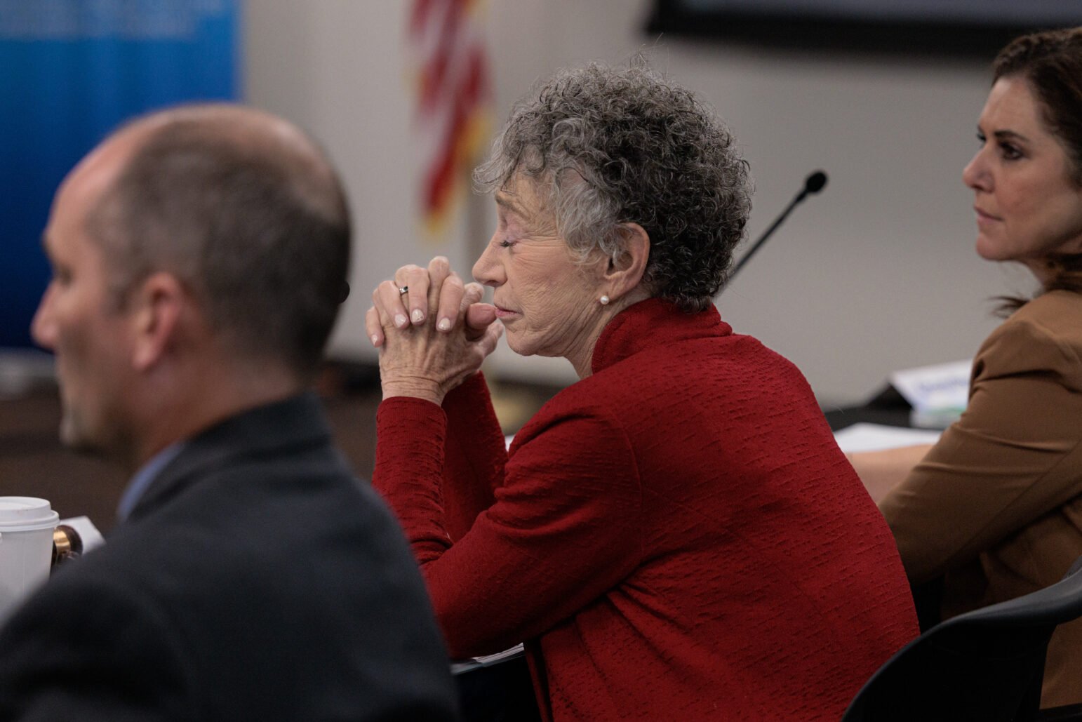 A woman in a red jacket, seated at a table with her hands clasped in front of her, appearing thoughtful during a meeting.