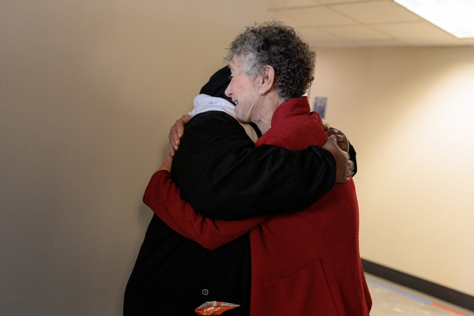 Two individuals hugging in a hallway, one in a red jacket, embracing a person in black.