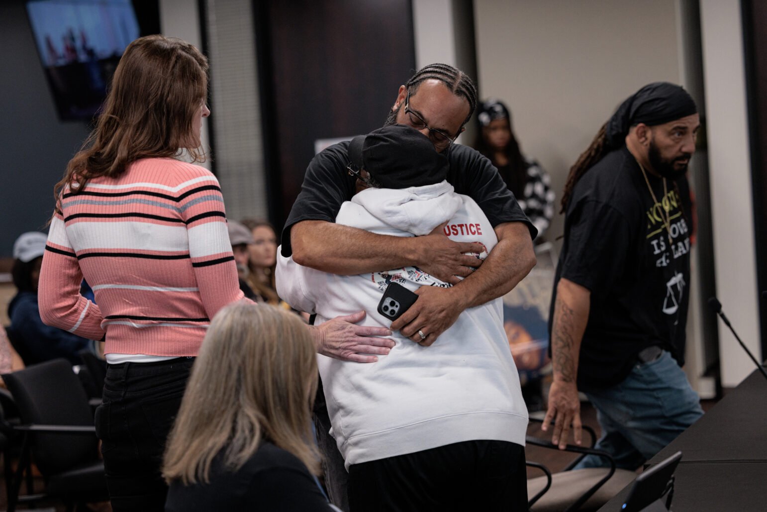 A man wearing glasses and a black shirt hugging a person wearing a white hoodie with the word "justice" on the back.