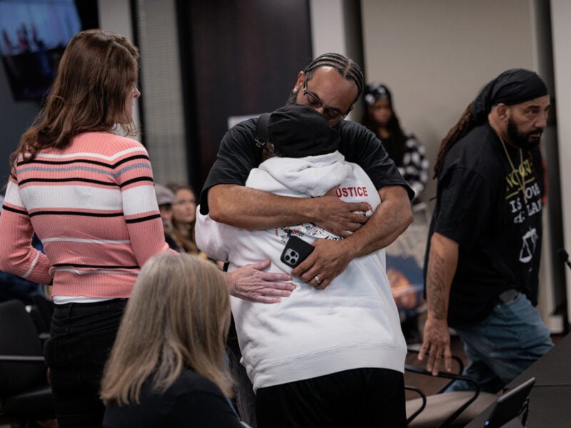 A man wearing glasses and a black shirt hugging a person wearing a white hoodie with the word "justice" on the back.