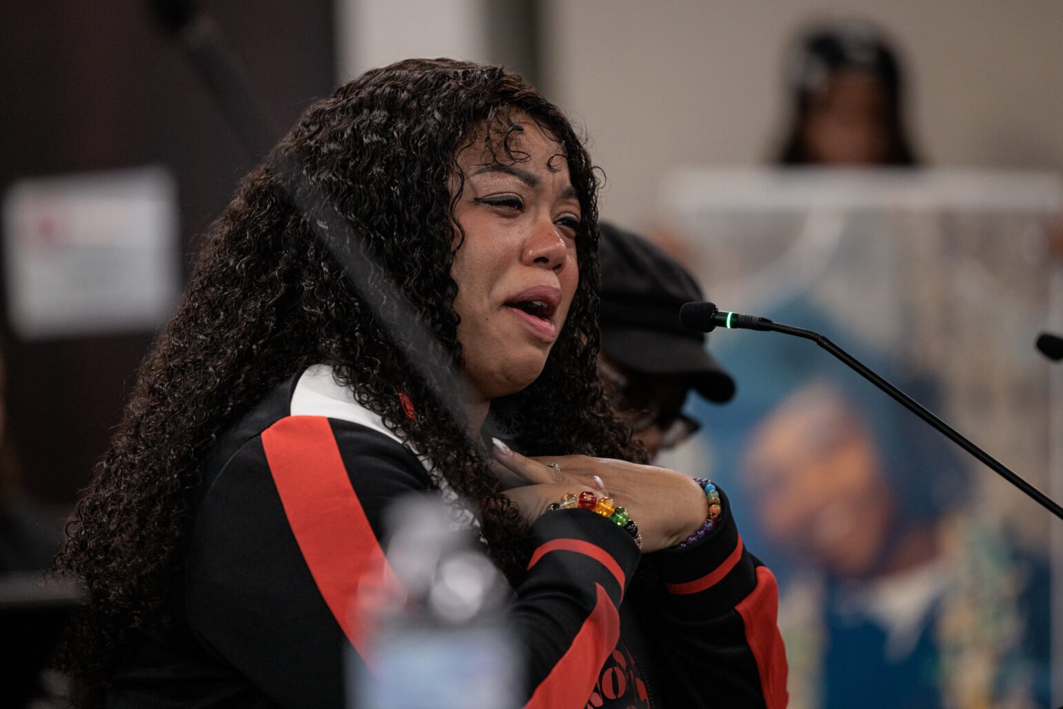 A woman speaking at a microphone, appearing emotional, wearing a black and red jacket with her hand on her chest.