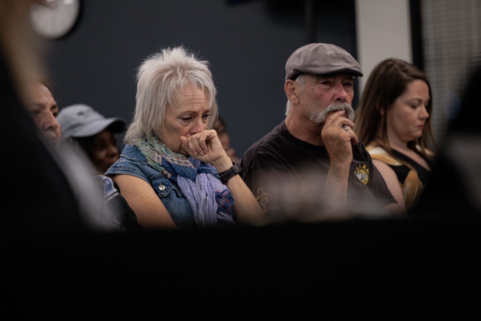 An older man and woman sitting in the audience of a meeting, both with their hands covering their mouths, appearing reflective or emotional.