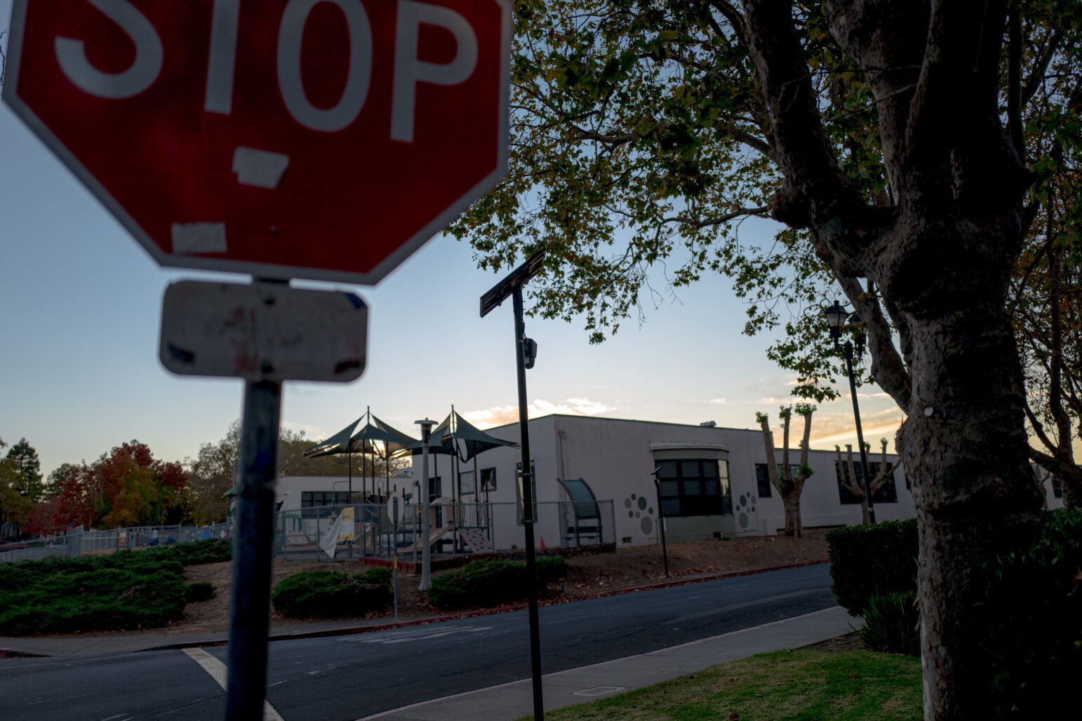 A stop sign in the foreground, partially obscuring the view of a playground and a light-colored building with trees lining the street. The evening sky is visible, with soft lighting and hints of sunset behind the scene. A surveillance device hangs from street sign in the center of the image.