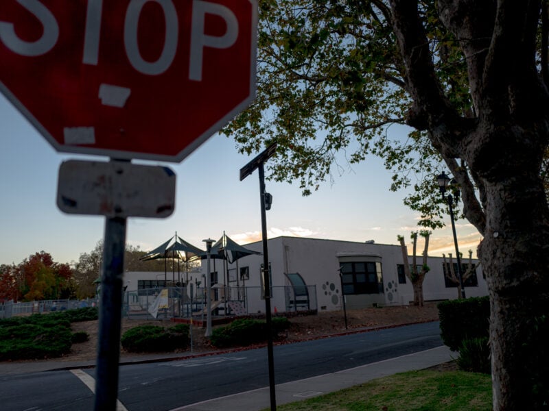 A stop sign in the foreground, partially obscuring the view of a playground and a light-colored building with trees lining the street. The evening sky is visible, with soft lighting and hints of sunset behind the scene. A surveillance device hangs from street sign in the center of the image.