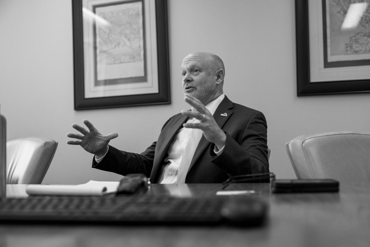 A black-and-white image of Andrew Murray sitting at a conference table. His hands are raised in a gesture, as he appears to be speaking or explaining something. He is seated in a formal office environment.