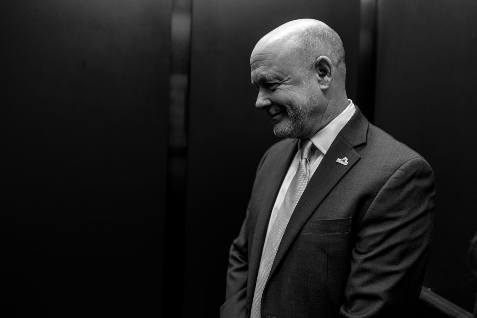 A candid black-and-white shot of Andrew Murray in an elevator, wearing a suit and tie, smiling and looking downward with a relaxed posture.