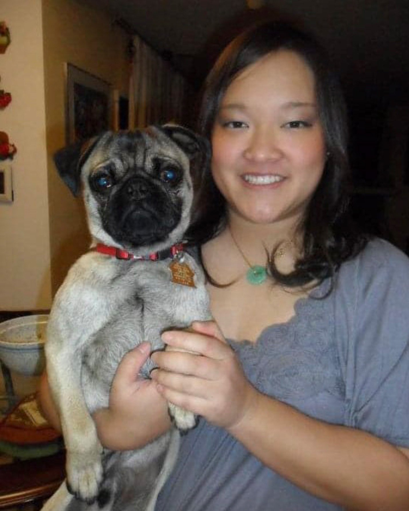 A woman smiles at the camera while holding a pug. The woman has long, dark hair and wears a light gray top with a green pendant necklace. The pug, wearing a red collar, looks directly into the camera with a neutral expression. They appear to be indoors, with household items visible in the background, giving the image a warm, casual atmosphere.