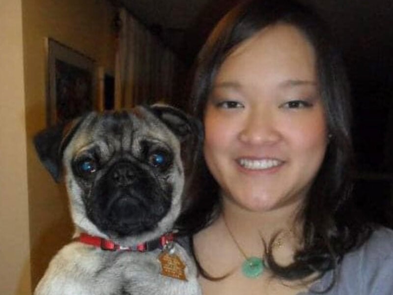 A woman smiles at the camera while holding a pug. The woman has long, dark hair and wears a light gray top with a green pendant necklace. The pug, wearing a red collar, looks directly into the camera with a neutral expression. They appear to be indoors, with household items visible in the background, giving the image a warm, casual atmosphere.