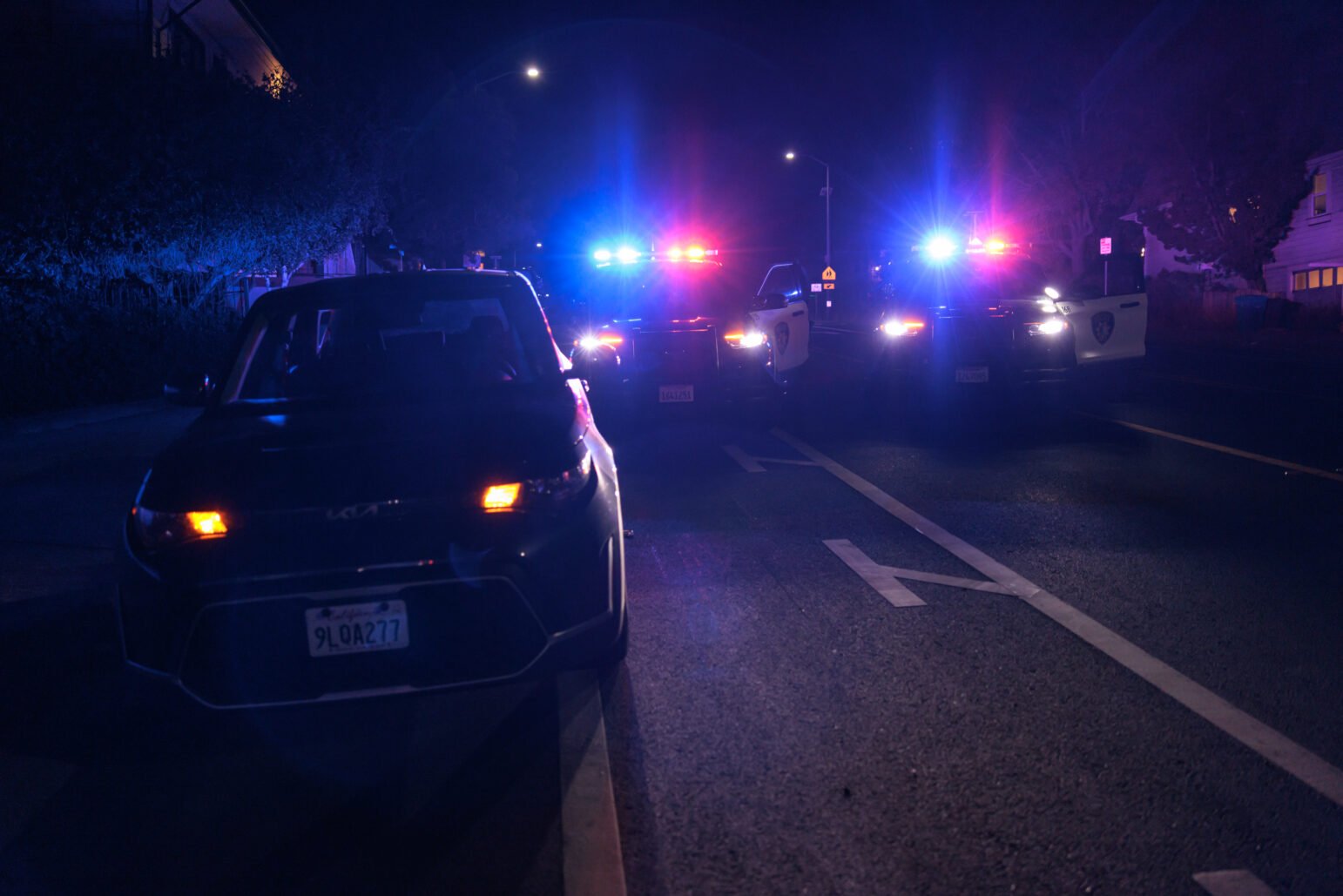 A nighttime scene showing a parked car with its hazard lights on, positioned in front of several police vehicles with flashing red and blue lights. The police cars illuminate the dark street, casting blue and red hues across the scene. The background is dimly lit, with faint outlines of buildings and trees.