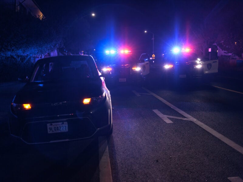 A nighttime scene showing a parked car with its hazard lights on, positioned in front of several police vehicles with flashing red and blue lights. The police cars illuminate the dark street, casting blue and red hues across the scene. The background is dimly lit, with faint outlines of buildings and trees.