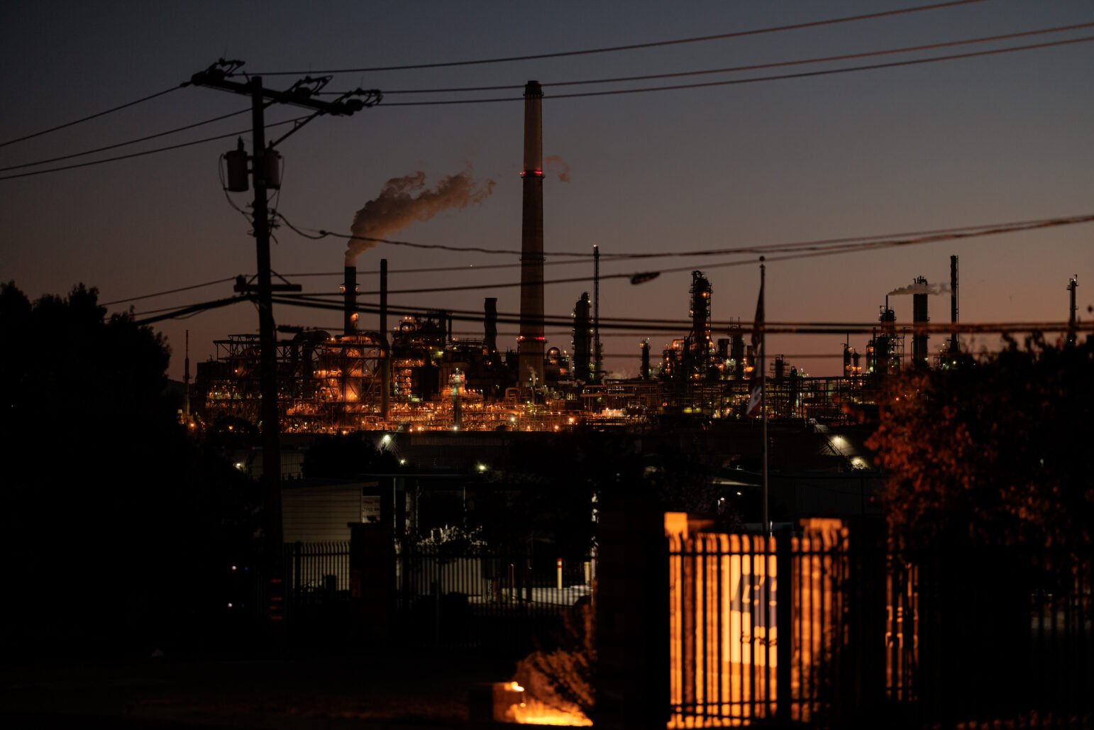 A nighttime view of an oil refinery illuminated by industrial lights, with emissions rising from a tall smokestack. The refinery’s structures are silhouetted against the twilight sky, with power lines and utility poles in the foreground adding to the industrial atmosphere.