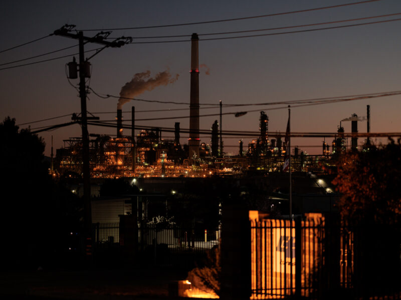 A nighttime view of an oil refinery illuminated by industrial lights, with emissions rising from a tall smokestack. The refinery’s structures are silhouetted against the twilight sky, with power lines and utility poles in the foreground adding to the industrial atmosphere.