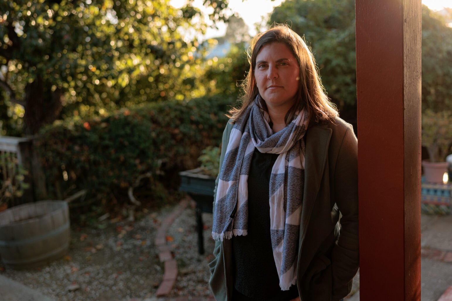 A woman stands outside near a wooden post, wearing a striped scarf and green jacket. The background shows a garden with soft, natural lighting.