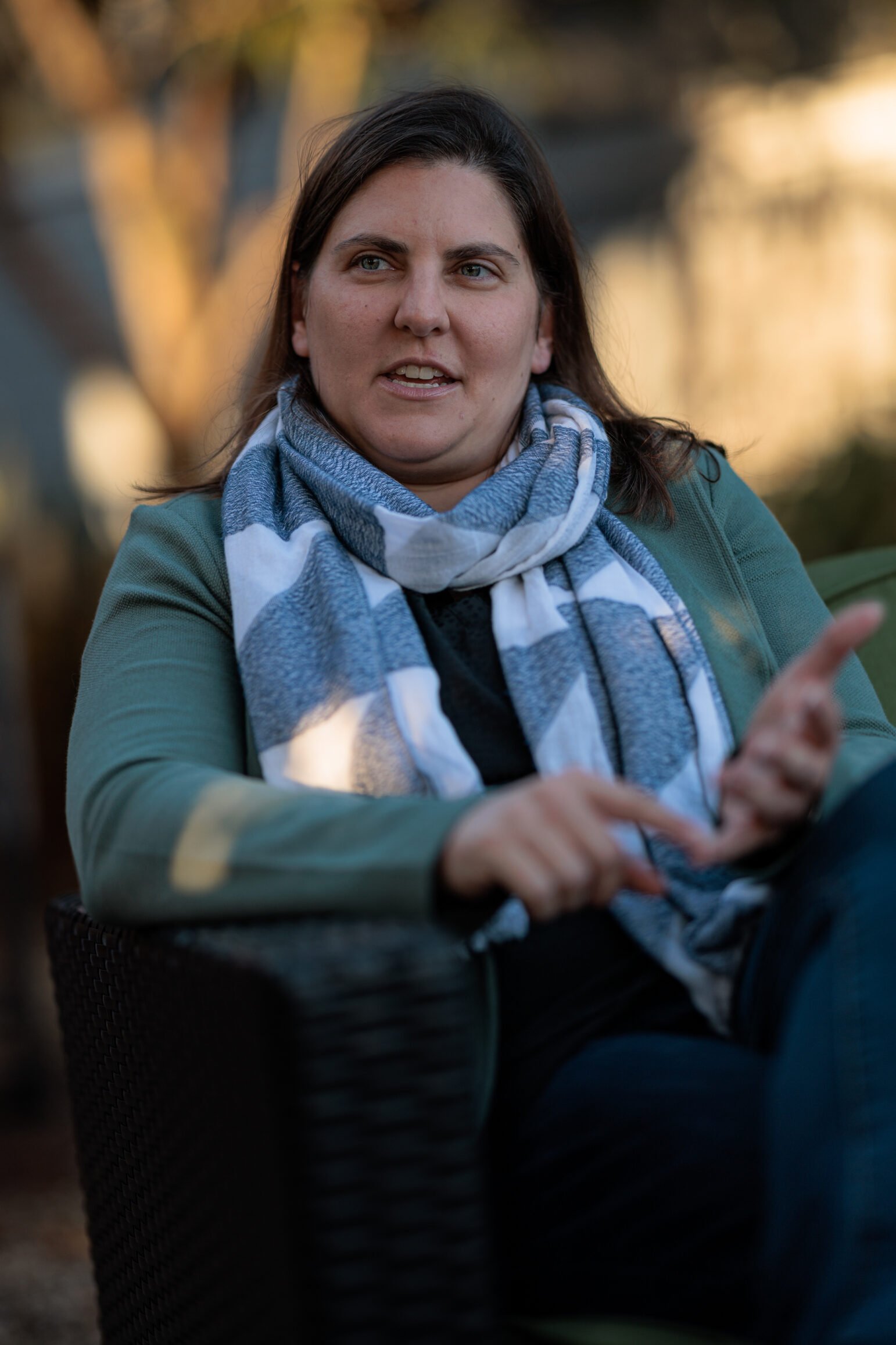 A woman sits in an outdoor chair, wearing a striped scarf and green jacket, speaking with a focused expression. The background is softly blurred.