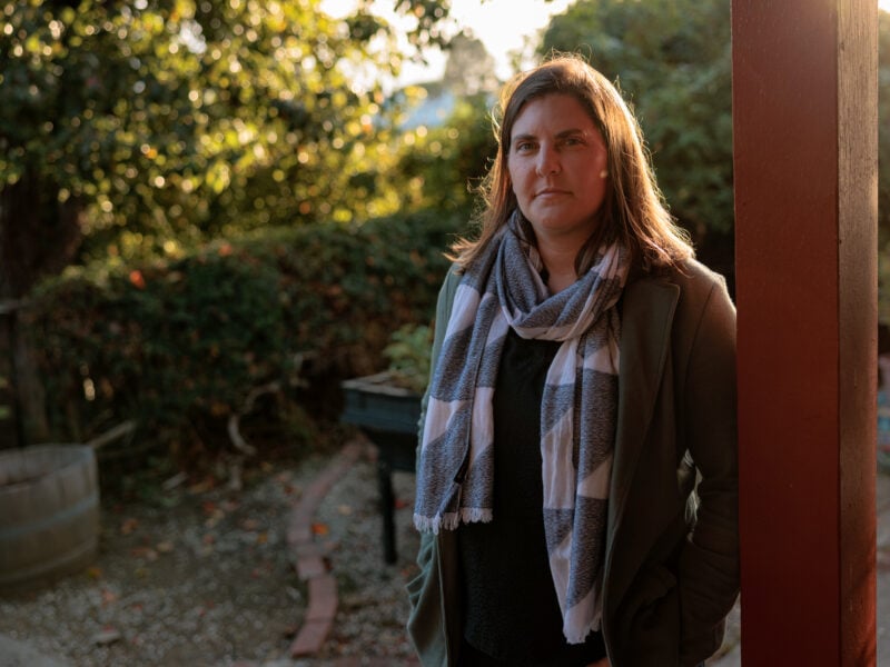 A woman stands outside near a wooden post, wearing a striped scarf and green jacket. The background shows a garden with soft, natural lighting.