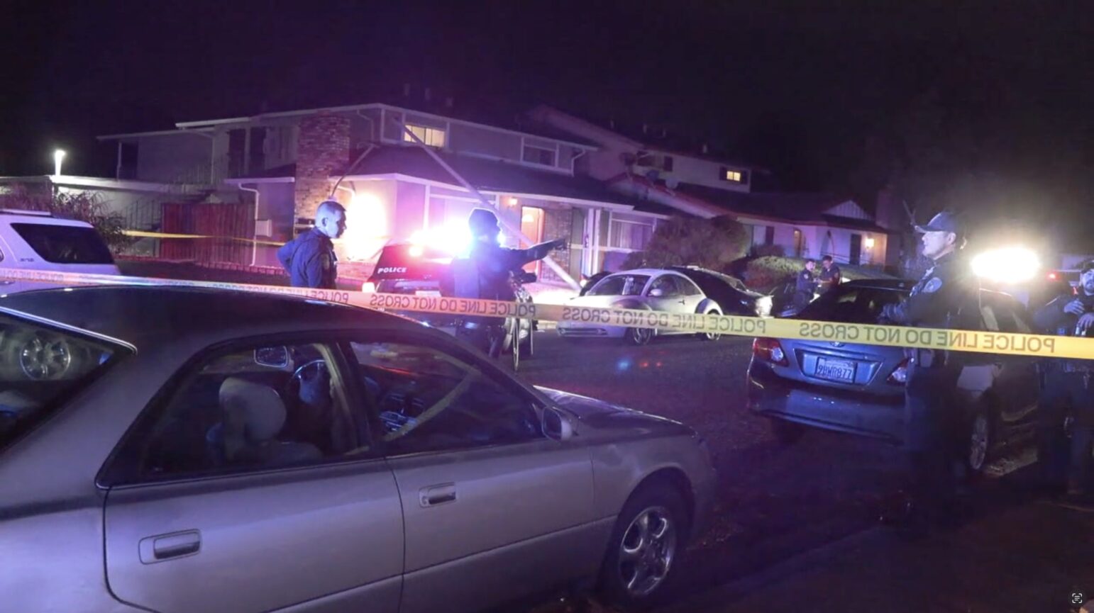 Police officers stand at a residential crime scene at night, surrounded by yellow police tape, with flashing emergency lights illuminating the area and parked vehicles visible in the foreground.