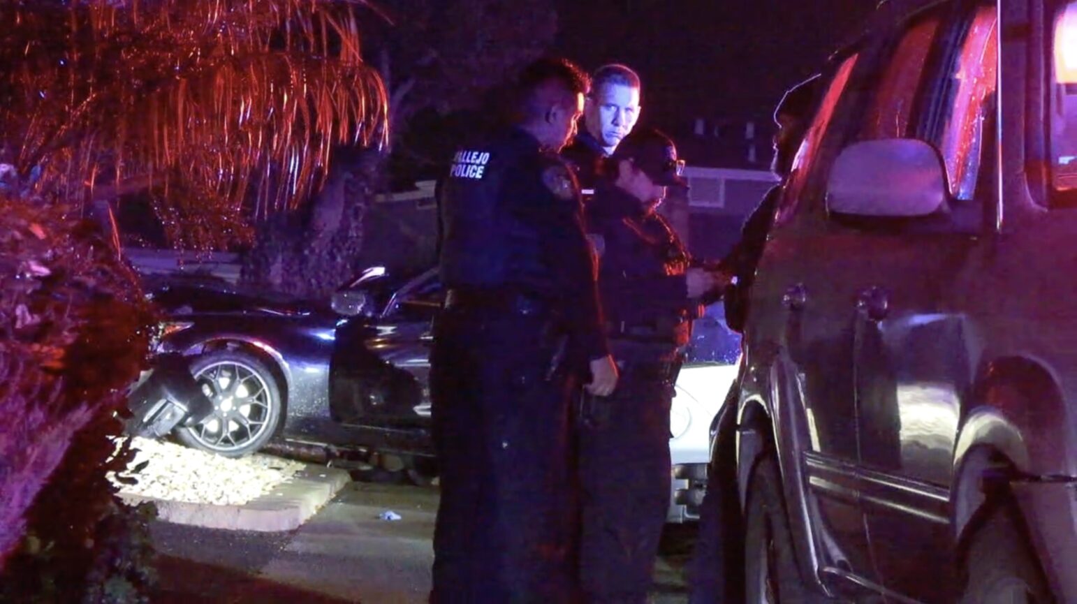 Vallejo police officers confer beside a damaged vehicle at a nighttime crime scene, with flashing red and blue lights reflecting off the surroundings and a tree in the background.