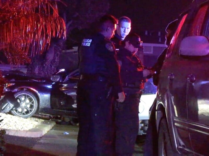 Vallejo police officers confer beside a damaged vehicle at a nighttime crime scene, with flashing red and blue lights reflecting off the surroundings and a tree in the background.