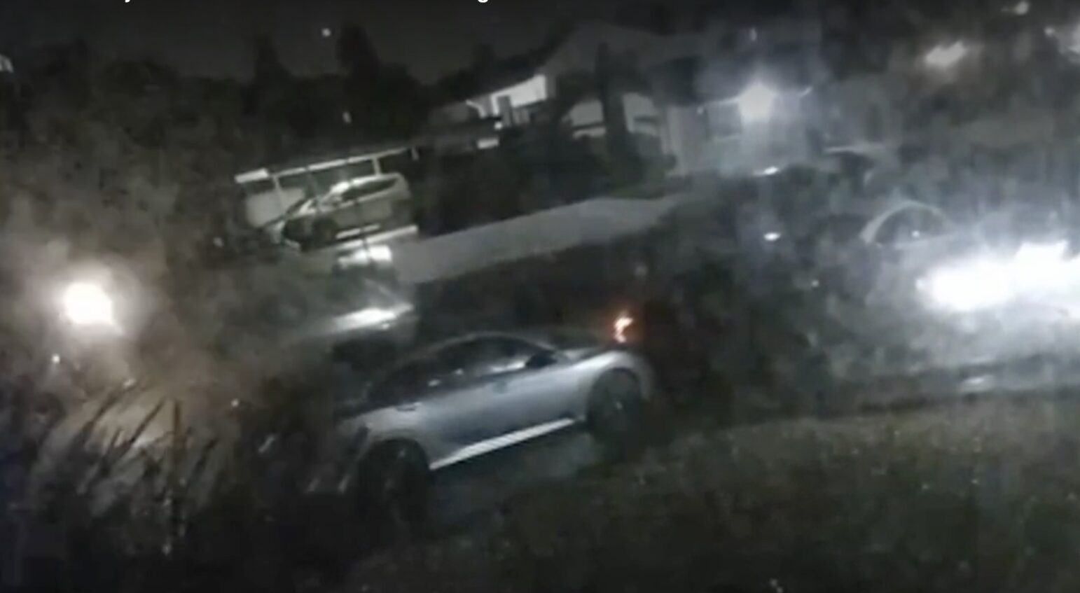 A grainy, nighttime surveillance image showing a street with a silver car in the foreground and additional vehicles and a house in the background, illuminated by streetlights and headlights.