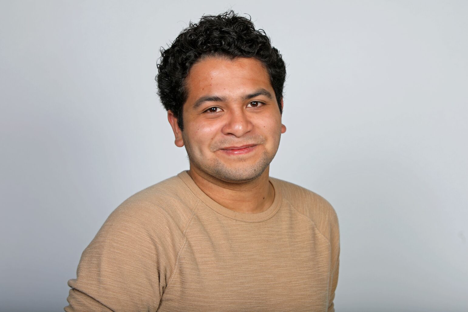 A portrait of a man with short, curly hair, wearing a light sweater and smiling softly against a plain background.