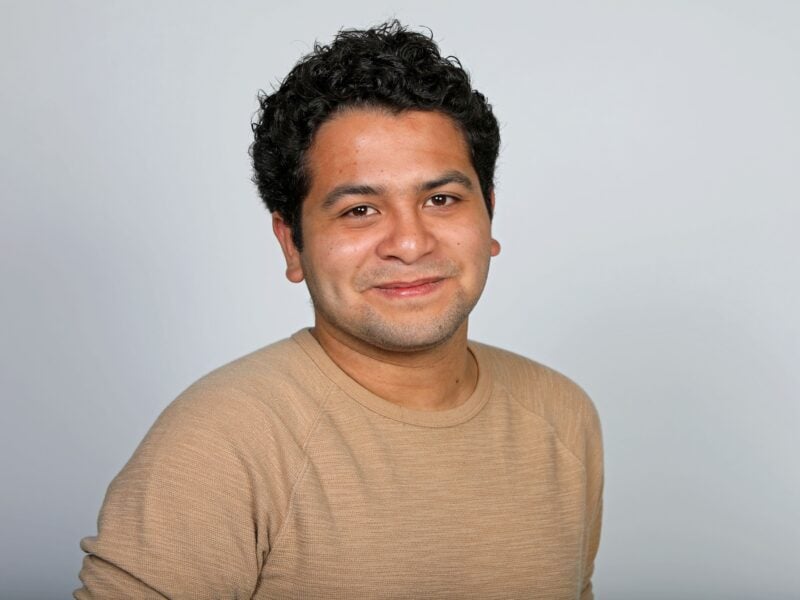 A portrait of a man with short, curly hair, wearing a light sweater and smiling softly against a plain background.